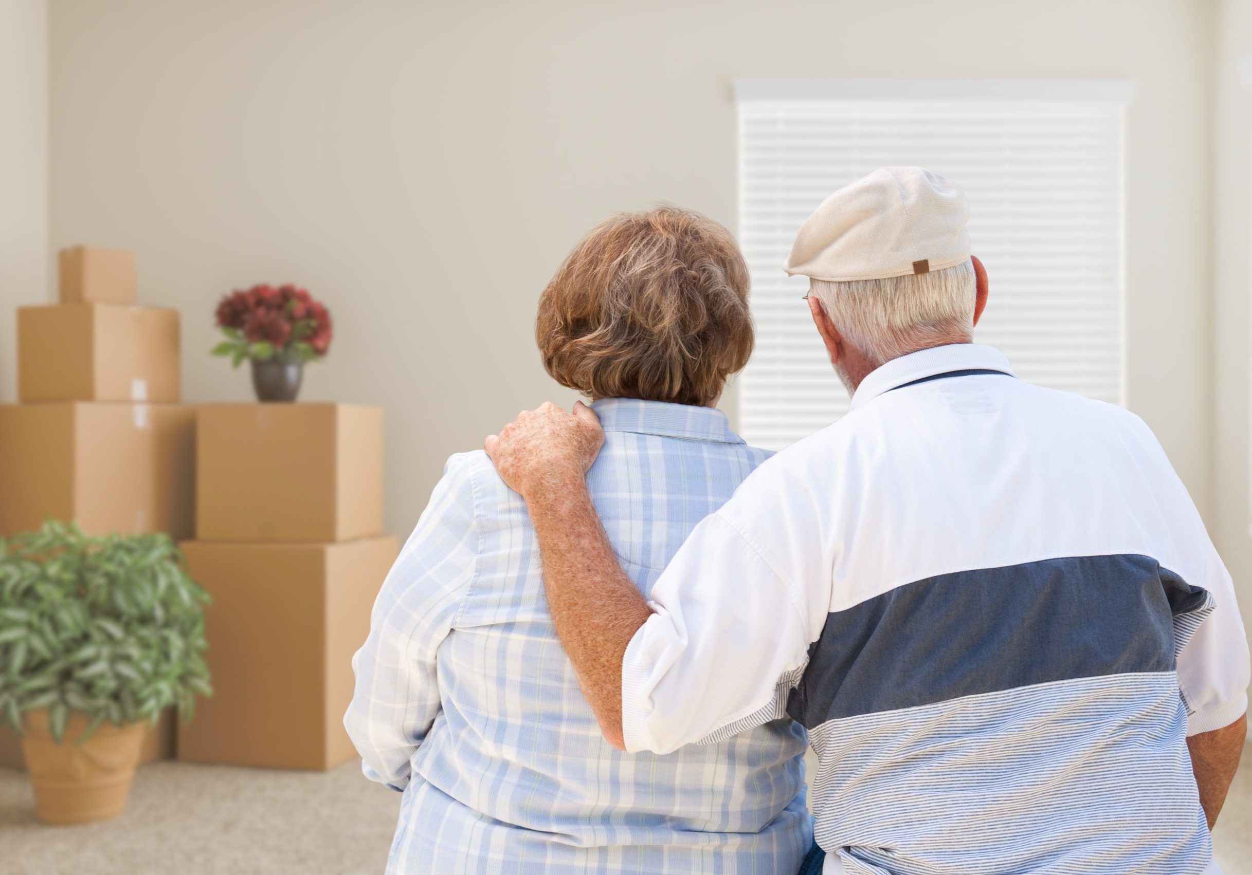 Senior Couple Facing Empty Room with Packed Moving Boxes and Potted Plants.